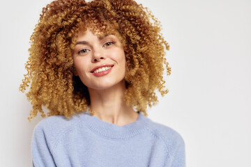Smiling woman with curly hair in a light blue sweater against a neutral background, showcasing a...