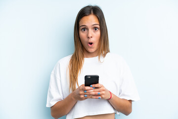 Young caucasian woman isolated on blue background looking at the camera while using the mobile with surprised expression