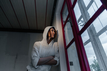 Pensive young caucasian female in hoodie looking out red-trimmed window on overcast day