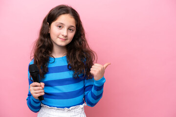 Little singer girl picking up a microphone isolated on pink background pointing to the side to...