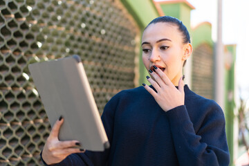 Young moroccan girl holding a tablet at outdoors with surprise and shocked facial expression