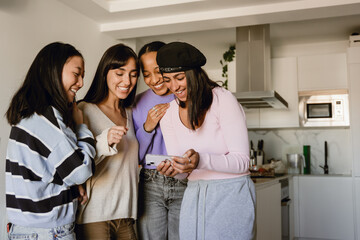 Obraz premium Four young women stand together in a modern kitchen, laughing and smiling while looking at a smartphone. They are enjoying a casual moment indoors during the daytime.