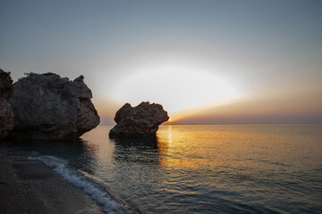 Landscape with a big rock on the beach near the sea. The sun is setting and the sky is orange
