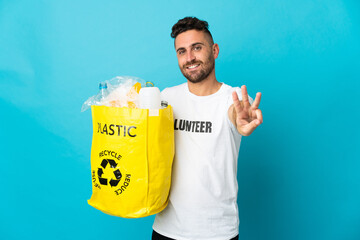 Caucasian man holding a bag full of plastic bottles to recycle isolated on blue background happy and counting three with fingers