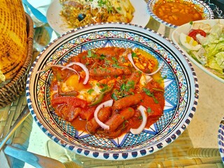 A vibrant and flavorful Tunisian dish called "Ojja" featuring spicy merguez sausages, poached eggs, bell peppers, onions, and tomato sauce, served in a traditional ceramic bowl.  © Hocineharoun