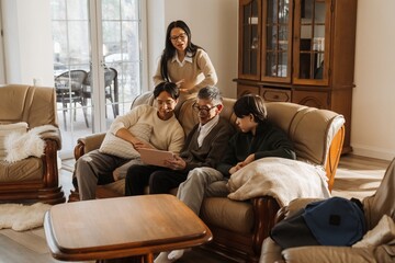 A senior Asian man sitting on a beige leather sofa indoors with his family, including two young Asian men and an adult Asian woman, smiling and looking at a tablet together in a cozy living room.