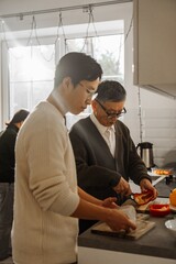 An elderly Asian man and a young Asian man prepare food together in a bright kitchen during the daytime. They cut vegetables and wear casual clothing.