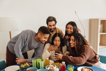 A diverse group of multiracial friends gather indoors around a table, looking at a smartphone and smiling. They sit together with snacks and drinks in a casual home setting.