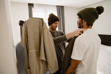 A young White man and a young Black man as a gay couple standing together in a bedroom, wearing knit beanies, choosing clothes from a hanger while getting ready near a bed and window.
