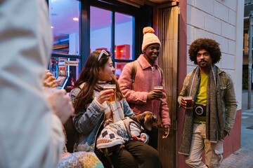 Several friends stand outside a bar, sipping beers and chatting while one woman holds a small dog in a sweater. The concept of evening meeting with friends in a city.
