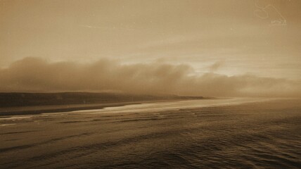 A sepia-toned seascape capturing a tranquil shoreline under soft, overcast skies.
