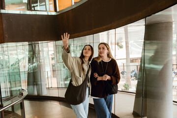 Two young White women walk arm in arm through a modern glass building, one of them pointing...