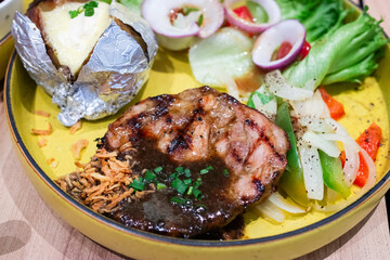 Grilled pork steak with black pepper sauce, served with baked potato, sautéed vegetables, and fresh salad on a yellow plate. A hearty and colorful Western-style meal.