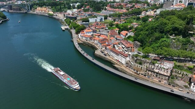 Aerial video of a river cruise ship navigating the Douro River near the riverside buildings and green hills of Porto, Portugal