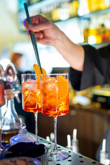 A bartender skillfully prepares two Aperol Spritz cocktails, garnished with orange slices, in a lively bar setting. The vibrant drinks in tall glasses filled with ice evoke a refreshing and social atm