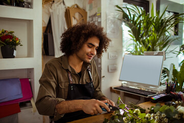 Young male florist arranging fresh flowers at work desk in flower shop