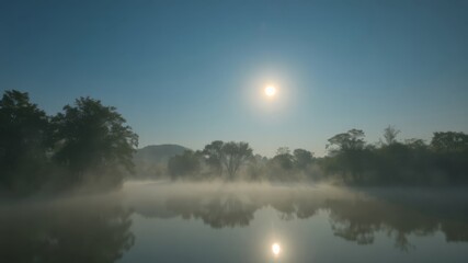 Fototapeta premium Serene morning mist rises over a calm lake reflecting the sun and trees creating a peaceful and tranquil landscape scene.