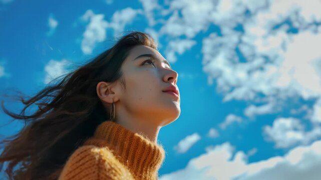 A woman with long hair is wearing a yellow sweater and looking up at the sky. The sky is blue with some clouds, and the woman is enjoying the view