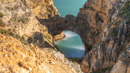 Blick von oben auf einen Felsenbogen an der Algarve, darunter Langzeitbelichtung des türkisen...