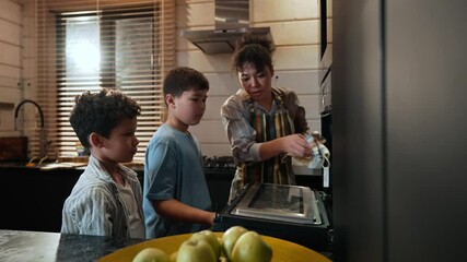 An adult, light brown-skinned mother and two school-aged sons put a pie in the oven to bake, then high-five each other and hug. A concept of family relationships, motherhood, and cooking.