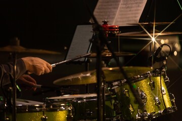 Close-up of a drummer&rsquo;s hands in action with a metallic green drum set under warm stage lights. Music sheets and microphones visible in the background. Copy space included.