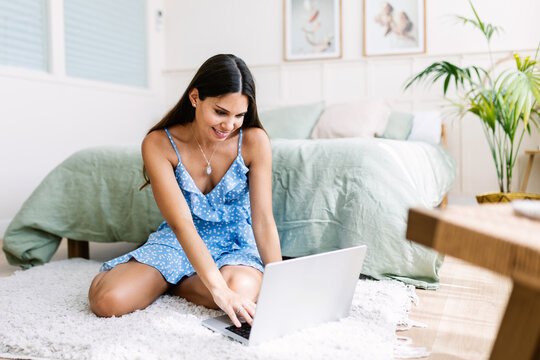 Smiling young woman sitting on a cozy carpet in her bedroom, working on her laptop and enjoying the comfort of remote work