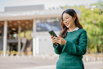 Fototapeta premium Asian female student in warm clothes using phone to chat with friends and ready for video call while in university.