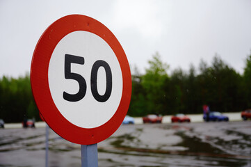 speed limit sign 50 km/h in focus with blurred racetrack and parked cars in the background. overcast day setting, emphasizing road safety and regulation imagery.