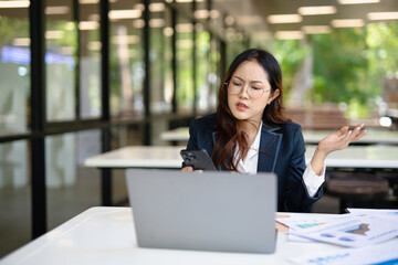 Fototapeta premium Asian businesswoman working on financial market research using laptop and digital tablet, stressed about digital investment in office.