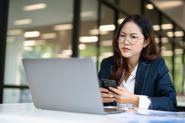 Asian businesswoman working on financial market research using laptop and digital tablet, stressed about digital investment in office.