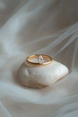 Delicate gold band ring with a single, pear-shaped clear gemstone rests atop a smooth, off-white stone, displayed on a sheer, ivory fabric