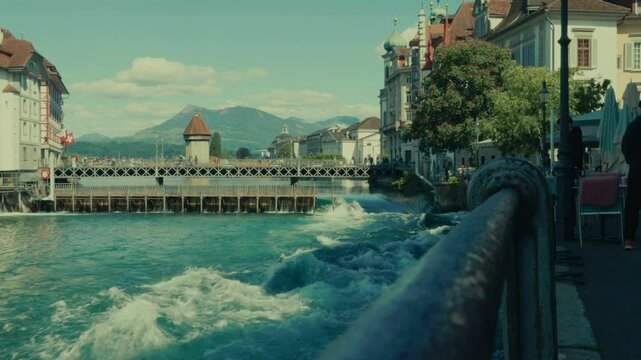 Die Kapellbr&uuml;cke in Luzern an einem sonnigen Tag &ndash; historische Holzbr&uuml;cke mit Wasserturm, gespiegelt im klaren See, umgeben von Alpen und blauem Himmel.