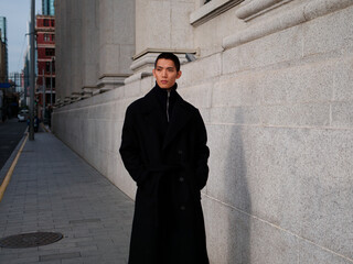 Portrait of handsome Chinese young man wearing black overcoat posing in the street, young guy with black short hair all in black with urban background. Male fashion, cool Asian young man lifestyle.