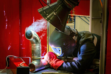 Welder in full protective gear working on metal pipe with visible smoke under extraction hood in...