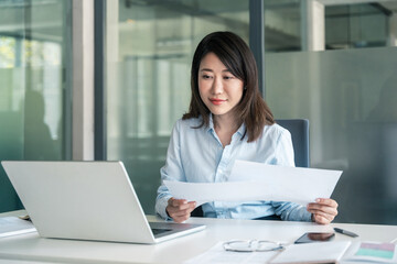 Focused Asian young business woman working on laptop computer reading financial document in office....
