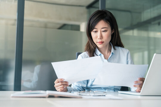 Thinking Asian young business woman working on laptop computer reading financial document in office. Accountant entrepreneur, banker manager businesswoman doing report documentation paperwork using pc
