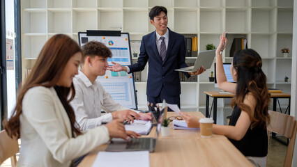 Team leader presenting data while female colleague raises her hand to participate during a meeting.