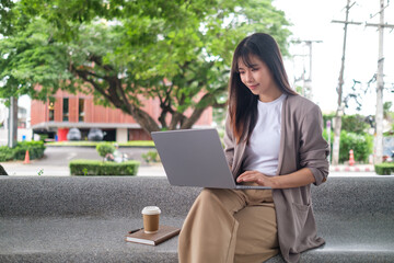 Woman in smart casual outfit using computer at a park bench, representing freelance or digital nomad lifestyle.