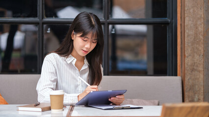 Fototapeta premium Professional Asian woman planning or working on a tablet with coffee and notes beside her.