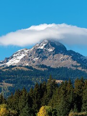 Fototapeta premium Snow-capped mountain peak with a fluffy cloud above, surrounded by forests under a clear blue sky.
