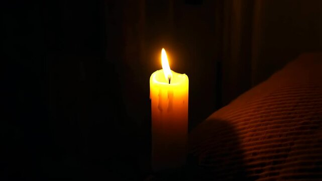 Close-up of glowing yellow candle flame lighting up dark bedroom next to rumpled bed sheets creating warm and cozy mood