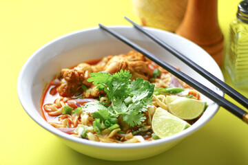 A bowl of spicy noodle soup with chicken, garnished with fresh cilantro, lime wedges, and chopped...