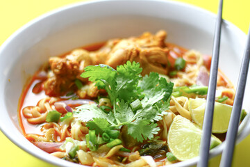 A bowl of spicy noodle soup with chicken, garnished with fresh cilantro, lime wedges, and chopped vegetables, served with black chopsticks. The vibrant colors and presentation are visually appealing.