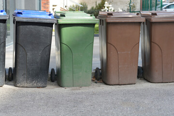 A row of wheeled garbage bins with different colored lids sits on an asphalt surface 
