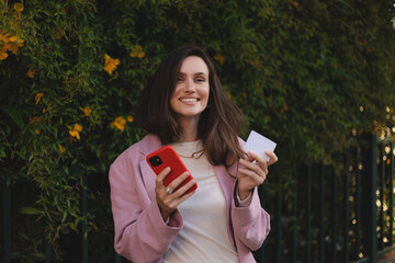 Cheerful young brunette woman shopping online with mobile phone and credit card while standing on a...