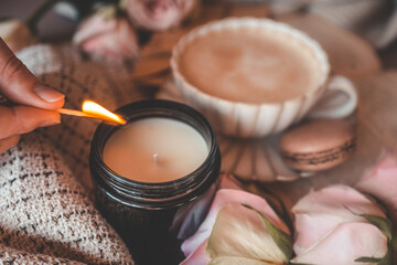 A cozy scene with a hand lighting a candle using a matchstick, surrounded by soft beige fabric, a cup of frothy coffee, pink roses, and a macaron. Warm and inviting atmosphere