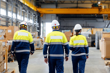 Group of warehouse workers wearing hard hats and reflective jackets uniform waking in industrial hangar of factory, back view