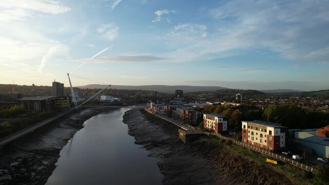Apartment houses on Usk river waterfront lit by setting sun. Backwards reveal of road bridge over river at low tide. Newport, Wales, UK