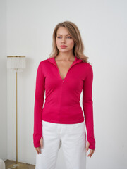 Woman in pink long-sleeve top with zipper, standing with neutral expression. She wears white pants....