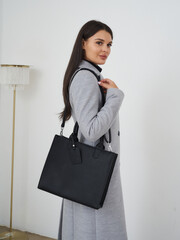 Young woman with long dark hair wearing a grey coat, holding a large black handbag. She is posing...